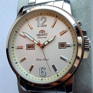 May include: Silver-toned Orient automatic watch with a white face. The watch features a date and day display, luminous hands, and a stainless steel band. The watch face displays the Orient logo, the number 12, and the number 6.