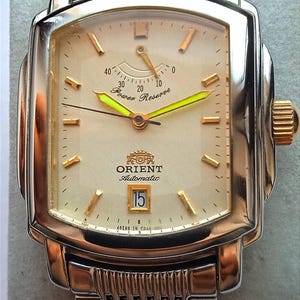 Pode incluir: Relógio de pulso automático Orient retangular com bracelete e caixa em metal prateado e dourado. O mostrador é creme com marcadores e ponteiros dourados e um indicador de reserva de marcha. A data é exibida às 6 horas.
