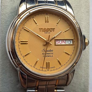 May include: A Tissot Seastar automatic watch with a gold-colored face and silver and gold-toned metal band. The watch face displays the day and date, with the text "SUN 13". The watch has a round face with gold hands and markers.