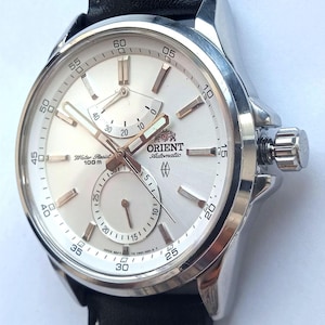 Puede incluir: Reloj Orient Automatic plateado con esfera blanca, subesferas y agujas/marcadores dorados. Correa de cuero negro con costuras blancas. La esfera muestra "Water Resist 100m". La caja del reloj mide unos 4 cm.