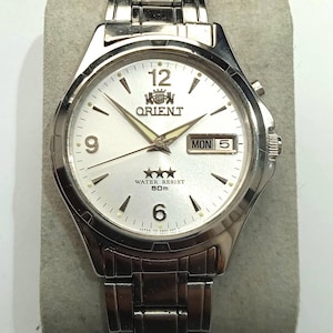 Può includere: Orologio Orient argentato con quadrante bianco, indici e lancette neri. Il quadrante mostra il giorno e la data, con la scritta "ORIENT" e "WATER RESIST 50m". L'orologio ha un cinturino argentato.