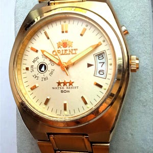May include: Gold-toned Orient watch with a cream-colored face. The watch features gold hands and markers, a day and date display, and the text "ORIENT" and "WATER RESIST 50m". The watch band is also gold-toned.