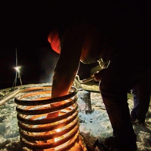 May include: A person in a red hat is using a tool to light a metal coil that is sitting on a snowy surface. The coil is glowing orange and red from the heat.