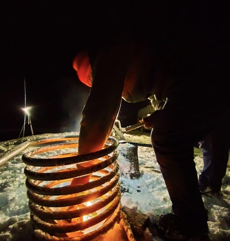 May include: A person in a red hat is using a tool to light a metal coil that is sitting on a snowy surface. The coil is glowing orange and red from the heat.