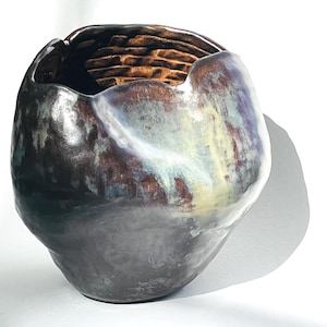 May include: A handcrafted ceramic vase with a unique, organic shape. The vase features a dark, iridescent glaze with shades of brown, blue, and green. The interior reveals a textured, brick-like pattern. The vase sits on a white surface.