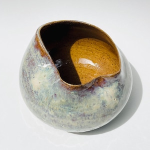 May include: A handcrafted ceramic bowl with a unique, asymmetrical shape. The exterior features a blend of blue, green, and purple hues, while the interior has a warm, brown glaze. The rim is accented with a darker brown. This decorative bowl is ideal for home decor.