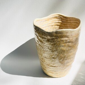 May include: A handmade ceramic vase with a textured, organic design. The vase has a light beige color with darker brown accents and a wavy rim. The exterior features a rough, layered texture, creating a unique, artisanal look. The vase casts a shadow on a white surface.