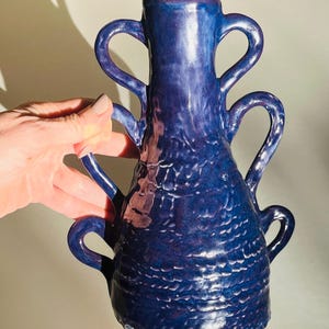 May include: A handmade, cobalt blue ceramic vase with four curved handles. The vase has a textured surface and a flared rim. The vase is approximately 10 inches tall.