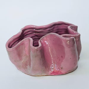 May include: A handcrafted, pink ceramic bowl with a wavy, ruffled design. The bowl has a glossy finish and a unique, organic shape. The interior features a textured surface, adding visual interest to this decorative piece.