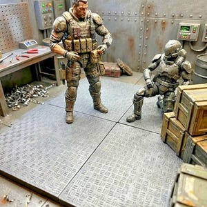 May include: Two detailed action figures in a diorama setting. One figure is a soldier in tan tactical gear standing upright, while the other is a futuristic armored soldier in a metallic gray suit kneeling on a textured gray floor panel.