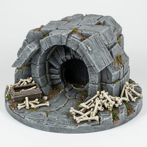 May include: A miniature stone dungeon entrance featuring a dark arched doorway, weathered grey masonry blocks, and scattered off-white skeletal remains. A small wooden crate sits near the entrance, with patches of green moss in the crevices.
