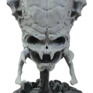 May include: A gray, detailed resin sculpture of a creature head with a prominent, ridged cranium, hollow eye sockets, and sharp fangs. The head is mounted on a central pillar rising from a dark, circular base covered in small, scattered skulls.