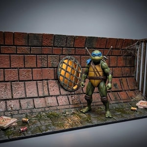 May include: A Teenage Mutant Ninja Turtle action figure stands in a diorama of a brick wall with a window and a metal grate. The figure is green and yellow, with a blue mask. Pizza boxes and cans are on the ground.