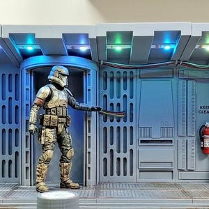 May include: A gray sci-fi diorama display featuring a central corridor with integrated blue and green LED ceiling lights. A detailed armored soldier figure stands in the doorway. A control console with warm lights and two small droid figures are present.