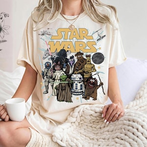 May include: A cream-colored t-shirt featuring a graphic print of Star Wars characters reimagined as Muppets. The design includes the yellow Star Wars logo, spaceships, and the Death Star, with characters like Kermit, Miss Piggy, and Fozzie Bear.