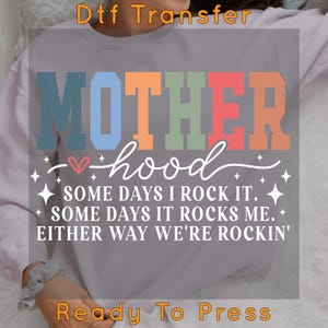 May include: A gray sweatshirt with a colorful graphic that reads "MOTHERhood" with a heart and stars. The text below reads "SOME DAYS I ROCK IT. SOME DAYS IT ROCKS ME. EITHER WAY WE'RE ROCKIN'." The text "Ready To Press" is at the bottom of the image.
