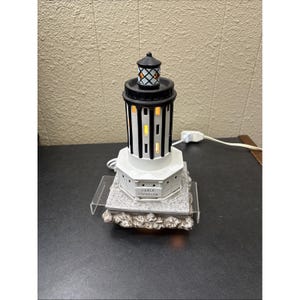 May include: A white and black lighthouse-shaped electric lamp with a black top and a base that reads "Cable Crossing". The lamp has a grid-like design and is lit from within, creating a warm glow. It sits on a textured base.