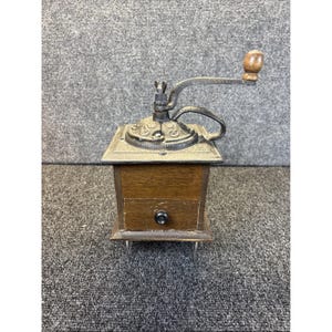 May include: A manual coffee grinder with a square wooden base, a small pull-out drawer featuring a black knob, and a cast iron top mechanism. The top includes an ornate metal plate, a curved handle arm, and a rounded wooden knob for grinding.