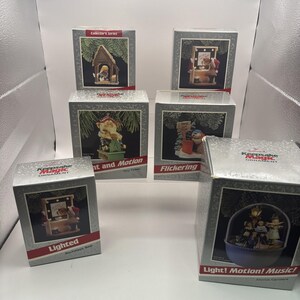 Puede incluir: Cinco cajas de adornos Hallmark Keepsake Magic con varias escenas navideñas. Las cajas muestran imágenes de un nacimiento, un oso en un tocador, un oso con un letrero, un oso en un taller y coristas. Cada caja incluye textos como Lighted, Flickering y Light! Motion! Music!