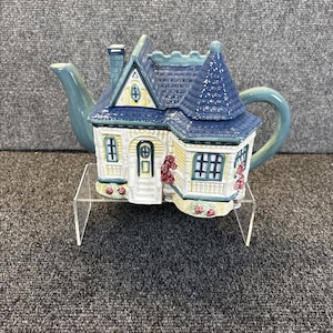 May include: A ceramic teapot shaped like a two-storey house with a blue shingled roof, a light yellow exterior, and white trim. The teapot features a blue spout, a curved blue handle, and small painted pink rose details around the base.