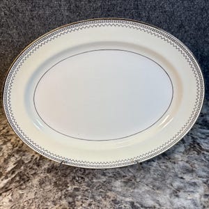 May include: An oval serving platter with a cream-colored surface and a decorative border. The border features a repeating geometric pattern in black and gold. A thin black line encircles the center of the platter.