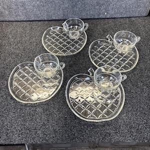 Könnte beinhalten: Vier klare Teetassen und Untertassen aus Glas. Die Untertassen sind wie Äpfel und Kreise geformt und haben ein Rautenmuster. Die Teetassen haben Henkel. Das Set steht auf einer dunkelgrauen Oberfläche.