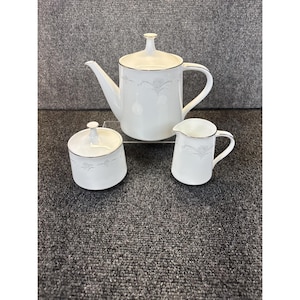 Może przedstawiać: Biały porcelanowy zestaw do herbaty z czajnikiem, dzbankiem do śmietanki i cukiernicą. Każdy element ma srebrne obramowanie i delikatny kwiatowy wzór. Czajnik ma długi dzióbek i zakrzywiony uchwyt.