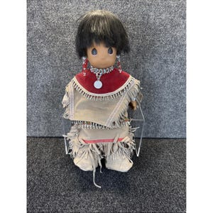 May include: A Precious Moments doll dressed in Native American-inspired attire. The doll has black hair and is wearing a red and tan outfit with fringe and beaded accents. The doll is wearing earrings and a necklace.