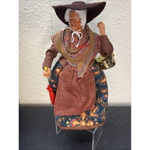 May include: A vintage figurine of a woman in traditional attire. She wears a large brown hat, a patterned shawl, and a red and orange checkered apron. The doll holds a small gold-colored container and a red pouch.