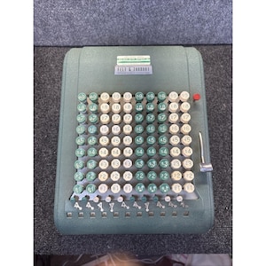 May include: A vintage sage green Felt & Tarrant Comptometer adding machine featuring a grid of alternating light cream and dark green circular keys labeled with numbers. A small red button sits at the top right, with a metal lever on the side.
