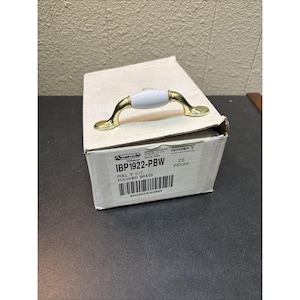 Puede incluir: Un tirador de armario de latón pulido con centro blanco, sobre una caja de cartón. El tirador mide 7,6 cm de centro a centro. La caja está etiquetada como "IBP1922-PBW" y "POLISHED BRASS".