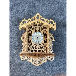 May include: A wooden wall clock with an ornate, carved design. The clock features a round face with Roman numerals and delicate hands. The clock is made of light-colored wood and has a small shelf at the bottom.