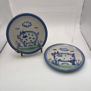 May include: Two round ceramic plates featuring a hand-painted blue pig with dark blue polka dots. The design includes a blue cloud, small birds, and green grass with orange flowers. The plates have a blue rim and a cream-colored background.