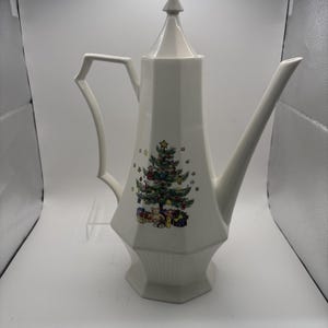 May include: A tall, white, hexagonal ceramic coffee pot featuring a pointed lid, a long angled spout, and a geometric handle. The front of the pot is decorated with a colorful illustration of a decorated Christmas tree with gifts underneath.