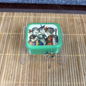 Puede incluir: Una pequeña caja de baratijas rectangular verde con tapa con bisagras. La tapa presenta una colorida ilustración de tres niños. La caja tiene un anillo de metal plateado adjunto en la parte inferior. La caja se exhibe en un soporte de plástico transparente.