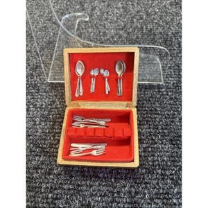 May include: A miniature silverware set in a wooden box with a red velvet interior. The set includes tiny spoons, forks, and knives, ideal for a dollhouse or miniature collection. The box is open, displaying the detailed silver utensils.