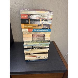 Può includere: Una pila di libri vintage con titoli visibili come "The Reason Why", "Delilah" e "The American Character". I libri hanno copertine colorate e sono impilati su una superficie scura, suggerendo una collezione di opere letterarie.