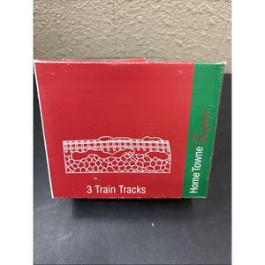 May include: A red and green rectangular box labelled Home Towne Express with the text 3 Train Tracks printed in white. The front of the box features a white line drawing of three train tracks resting on a stone-like base with snow accents.