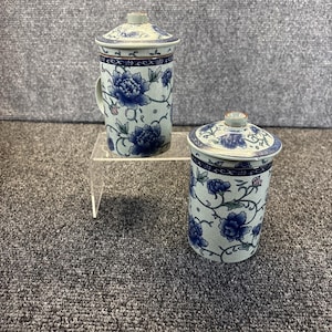 May include: Two ceramic tea mugs with lids, featuring a light blue base color and a dark blue floral pattern of peonies and vines. One mug includes a side handle, while the other is a straight-sided cylindrical vessel with a matching lid.