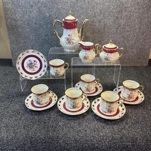 May include: A porcelain tea set featuring a teapot, creamer, sugar bowl, and four teacups with matching saucers. Each piece is cream-coloured with deep red bands, gold trim, and colourful floral bouquets. The saucers have intricate lattice-cut edges.