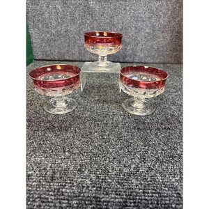 Puede incluir: Tres copas de postre de cristal vintage con bordes rojo rubí. Las copas tienen una base transparente con pie y un diseño decorativo grabado alrededor del cuenco. Las copas están dispuestas sobre una superficie gris.