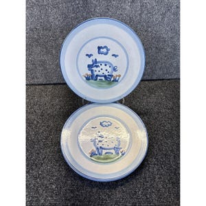 May include: Two ceramic plates with a blue and white design. Each plate features a central illustration of a pig with blue spots, set against a backdrop of grass, clouds, and birds. The plates have a blue border.