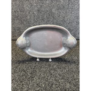 May include: Oval-shaped, silver-toned metal tray with decorative wheat sheaf designs on each end. The tray has a shallow, recessed center and rests on three small, ball-shaped feet. The overall design is vintage and elegant.