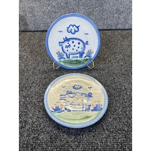 以下が含まれることがあります： 豚のデザインが施された2つのセラミックプレート。プレートは白い中心部と青い縁取りがあります。豚は白い斑点のある青色で、青い雲と緑の草があります。プレートは透明なスタンドにあります。