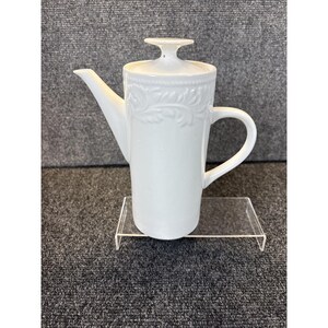 May include: A white ceramic coffee pot with a lid and a handle. The pot has a long spout and is decorated with a floral pattern on the upper portion. The pot is displayed on a clear acrylic stand.