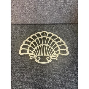 May include: A decorative, fan-shaped object with a scalloped edge and intricate cut-out design. The object is a light, neutral color and appears to be made of metal or a similar material. The design features a series of vertical lines and curved elements.