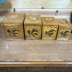May include: Set of four wooden canisters with rooster designs. The canisters are painted in shades of brown, yellow, and black. Each canister has a rooster painted on the front with a black, yellow, and white design.