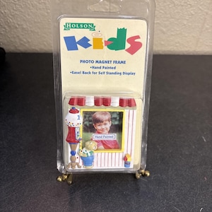 May include: A photo magnet frame for kids, featuring a hand-painted design. The frame has a red and white striped awning, a gumball machine, and a small bear. The frame is designed for self-standing display.