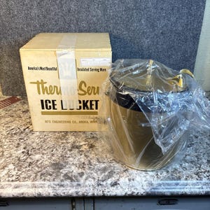 Puede incluir: Un cubo de hielo Thermo-Serv de color dorado con un asa negra, aún en su embalaje original. La caja dice "America's Most Beautiful" y "Insulated Serving Ware."