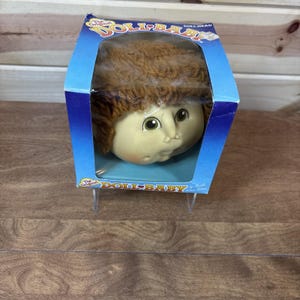 Op de afbeelding: Een poppenhoofd met bruin wollen haar en groene ogen, verpakt in een blauwe kartonnen doos met een doorzichtig plastic venster. De verpakking bevat de tekst The Original DOLL BABY en DOLL HEAD in witte en gele letters op het voorpaneel.